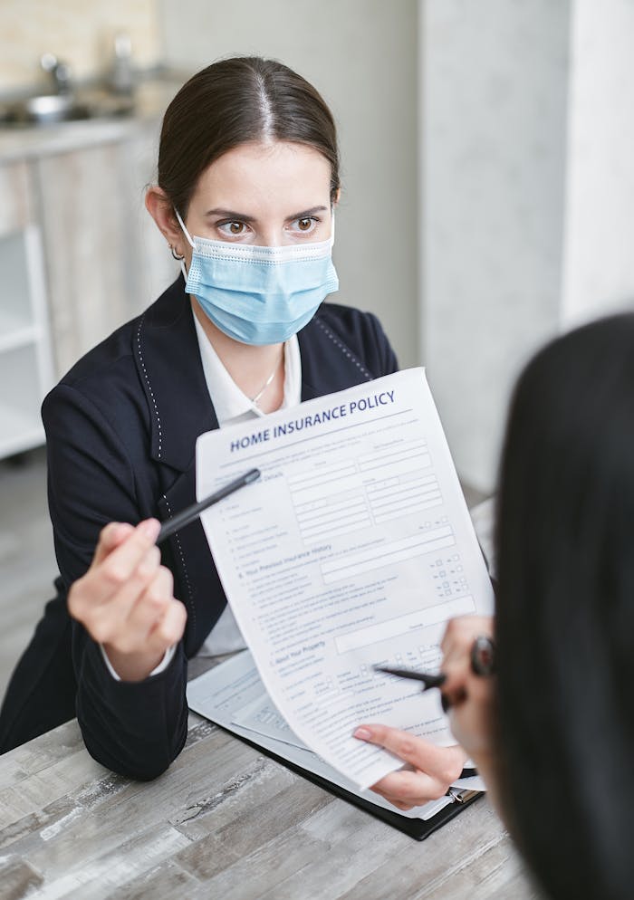 Professional woman wearing a face mask discussing a home insurance policy with a client at a desk.